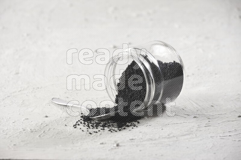 A glass jar and a metal spoon full of black seeds and the jar flipped and seeds spread on a textured white flooring