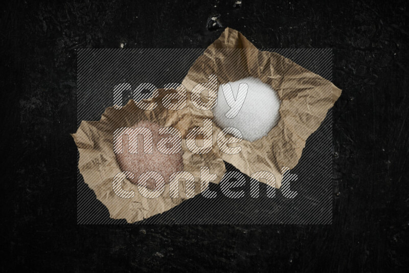 2 crumpled pieces of paper full of pink himalayan salt and white salt on black background