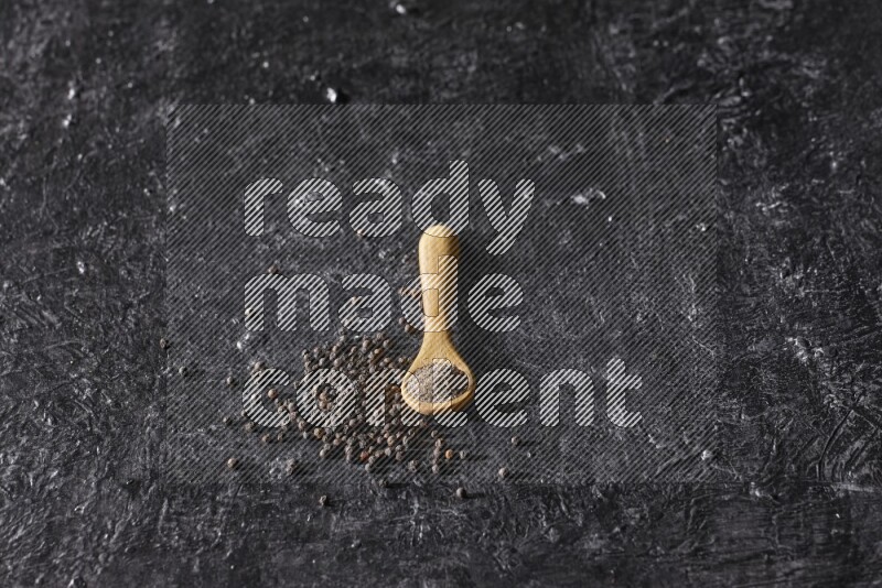 A wooden spoon full of black pepper powder and black pepper beads spread on a textured black flooring
