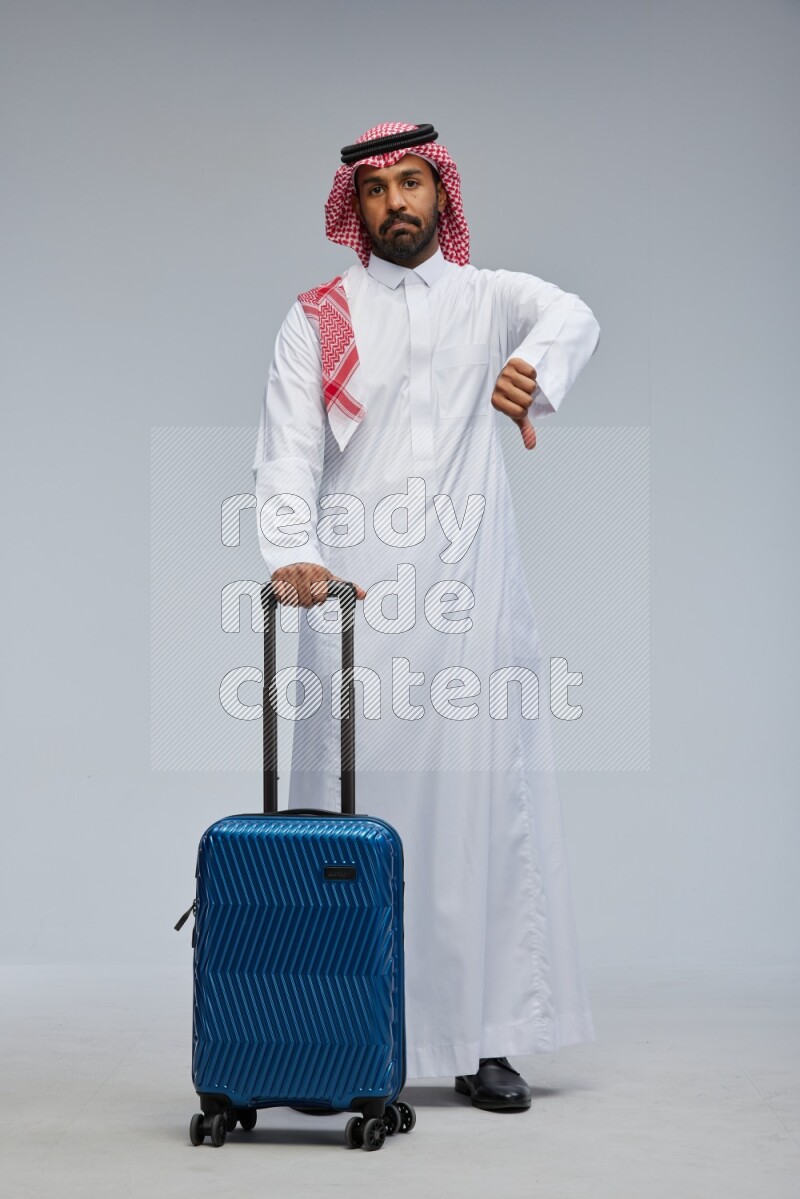 Saudi man wearing Thob and shomag standing holding Travel bag on Gray background