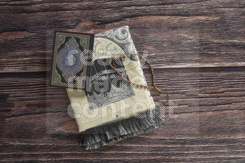 A folded prayer mat with different elements such as quran and prayer beads on brown wooden background