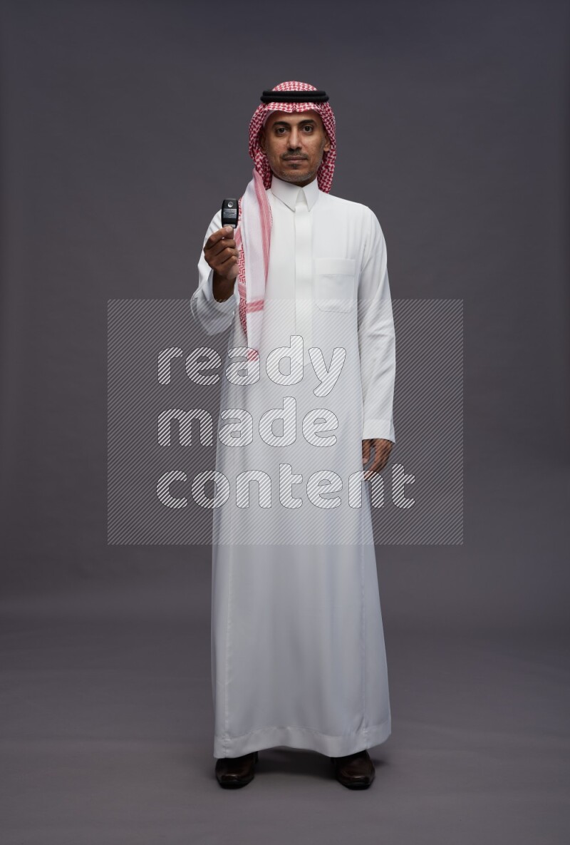 Saudi man wearing thob and shomag standing holding key car on gray background