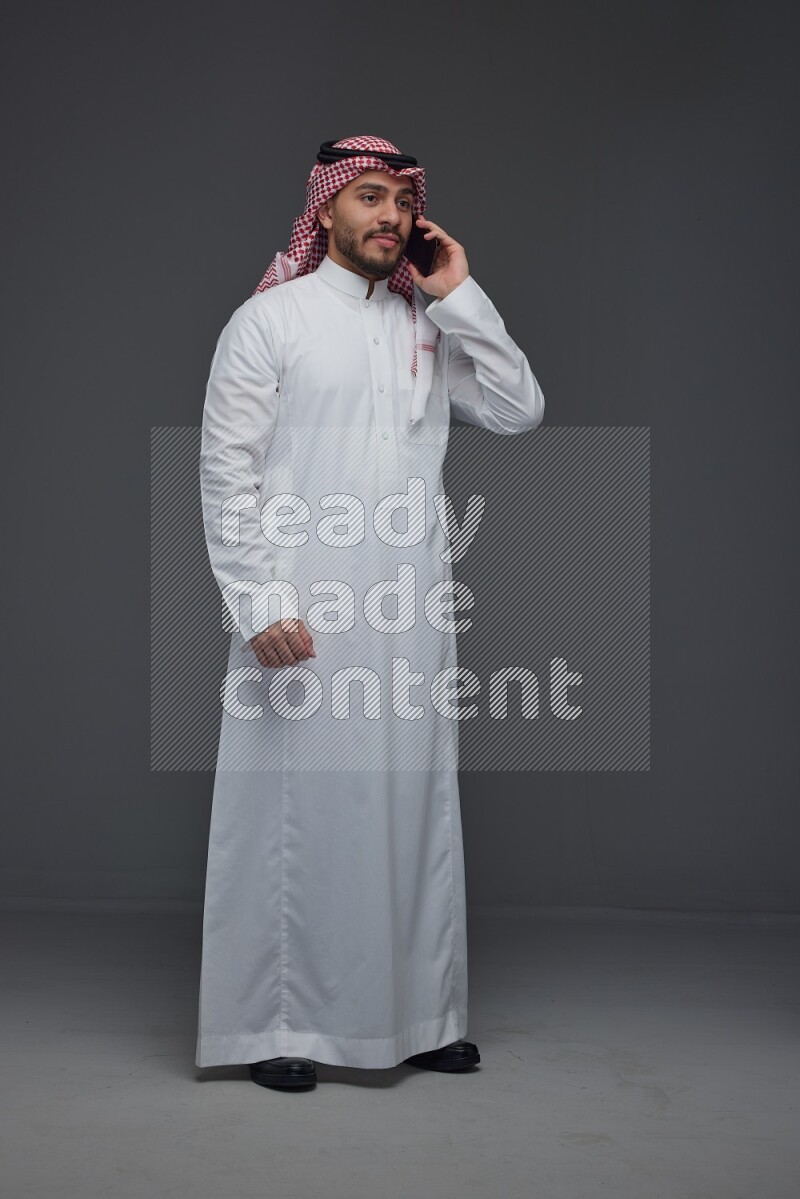A Saudi man wearing Thobe and Shmagh talking in the phone eye level on a gray background