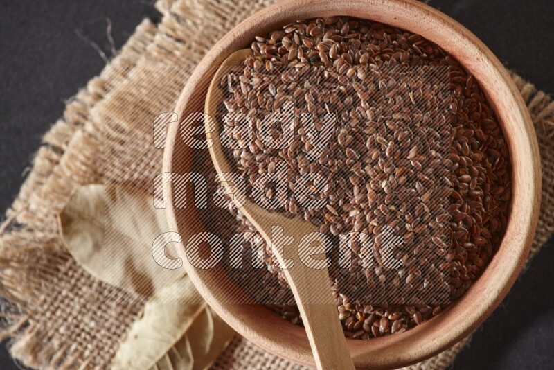A wooden bowl full of flaxseeds with wooden spoon full of the seeds on it on burlap fabric on a black flooring