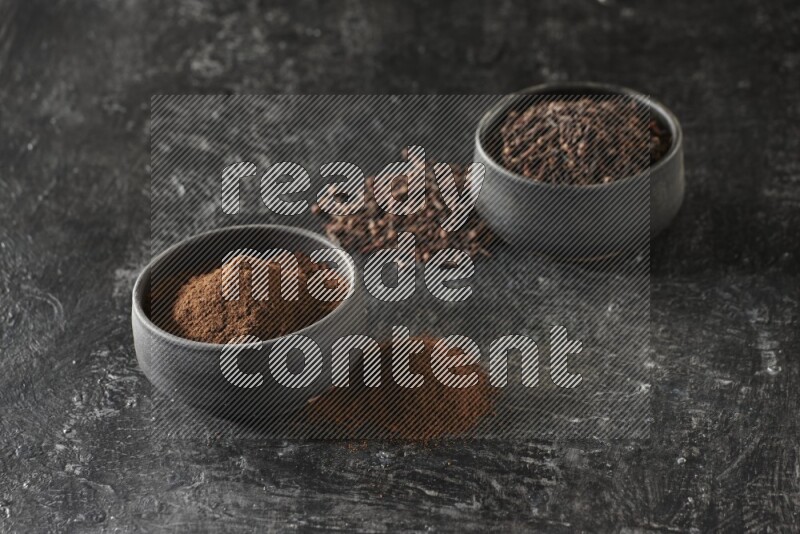 2 Black pottery bowls full of cloves and the other full of cloves powder on textured black flooring