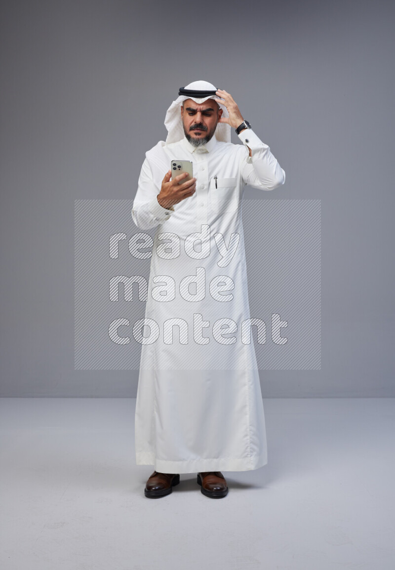 Saudi man Wearing Thob and white Shomag standing texting on phone on Gray background
