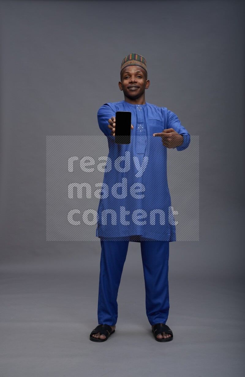 Man wearing Nigerian outfit standing showing phone to camera on gray background