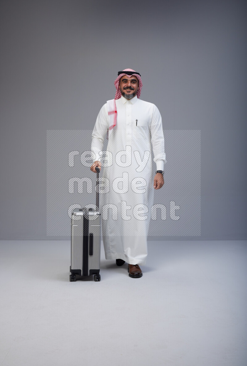 Saudi man wearing Thob and red Shomag standing holding Travel bag on Gray background