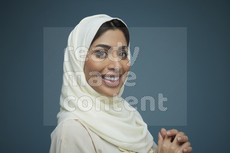 A Saudi woman posing in a blue background wearing an off-white Abaya with hijab