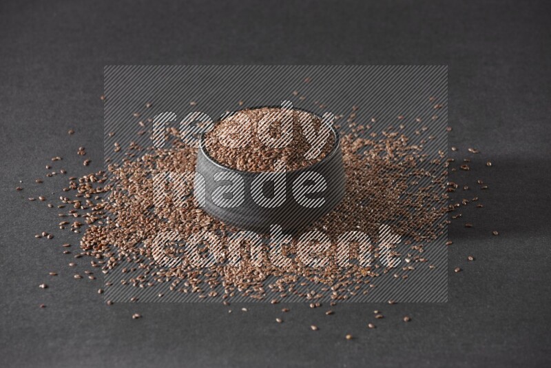 A black pottery bowl full of flaxseeds surrounded by the seeds on a black flooring