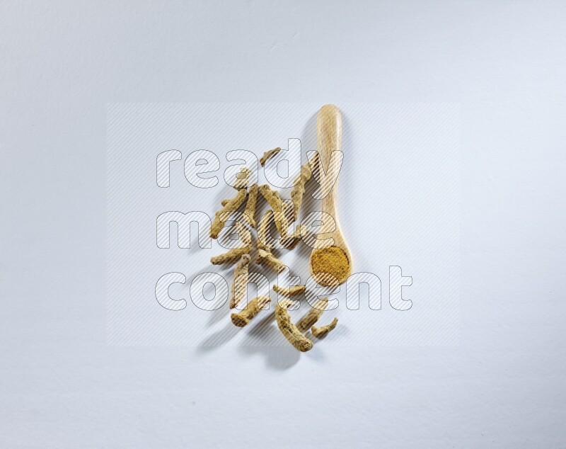 A wooden spoon full of turmeric powder with dried turmeric fingers beside it on white flooring