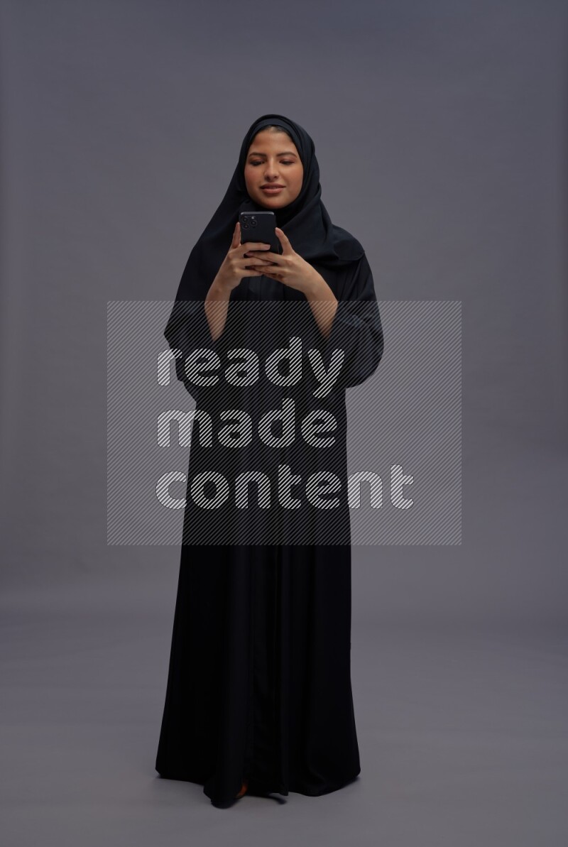 Saudi woman wearing Abaya standing texting on phone on gray background