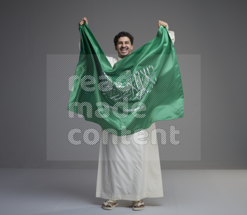 A Saudi man standing wearing thob holding big Saudi flag on gray background