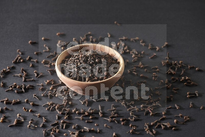 A wooden bowl full of cloves with spreaded whole cloves on a black flooring