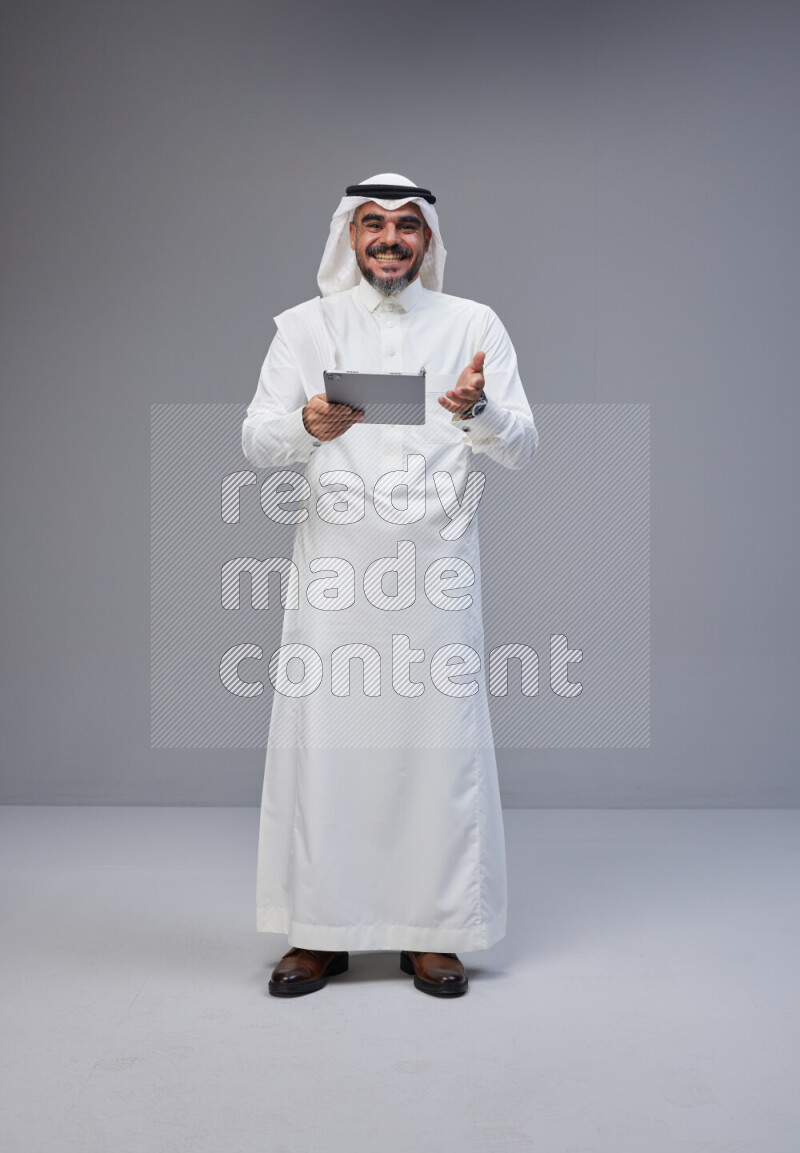 Saudi man Wearing Thob and white Shomag standing working on tablet on Gray background
