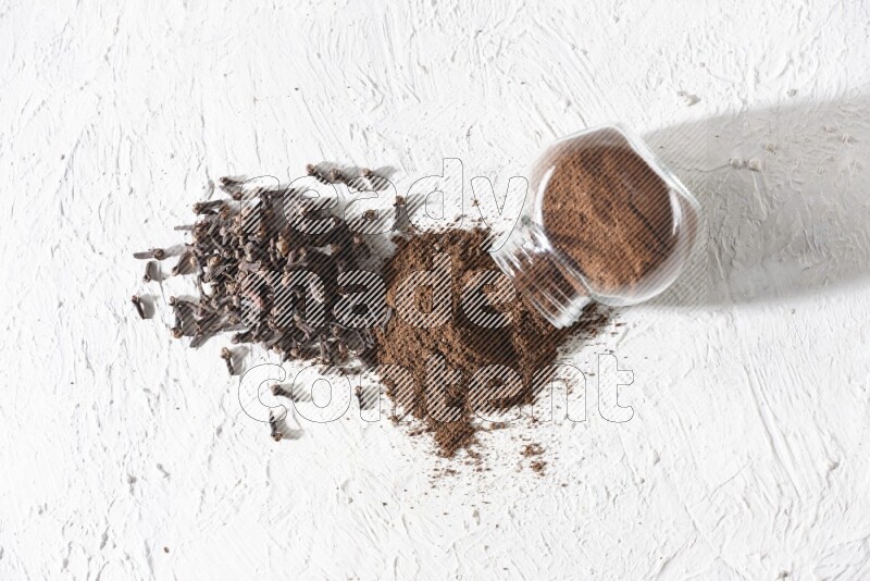 A glass spice jar full of cloves powder flipped and powder came out of it with cloves spread on textured white flooring