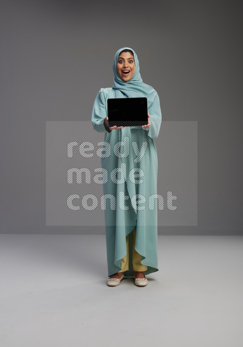 Saudi Woman wearing Abaya standing showing tablet to camera on Gray background