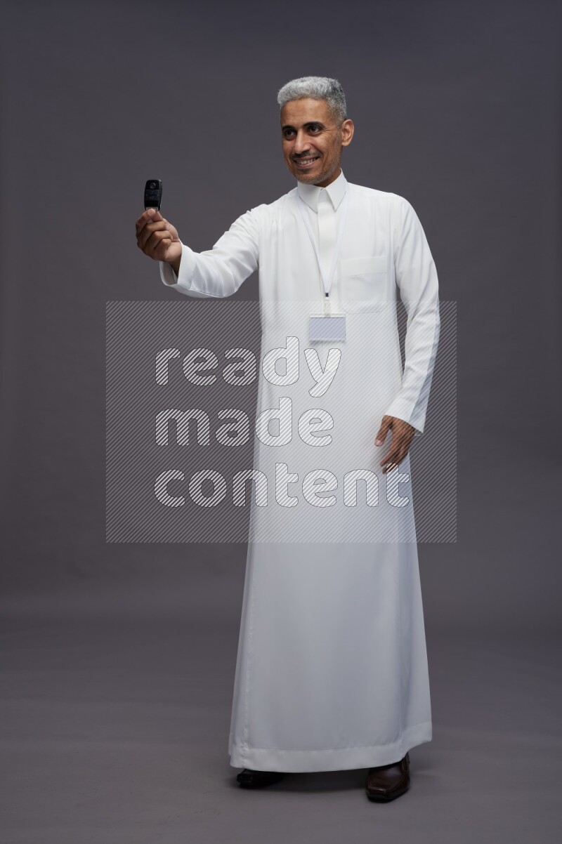 Saudi man wearing thob with neck strap employee badge standing holding key car on gray background