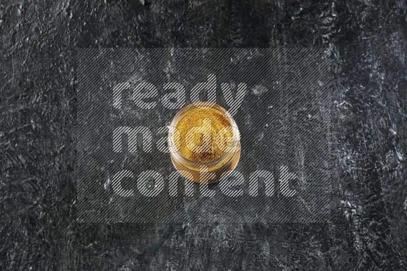 A glass jar full of turmeric powder on a textured black flooring