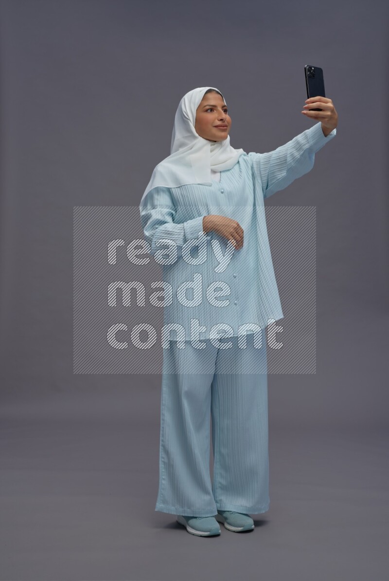 Saudi woman wearing hijab clothes standing taking selfie on gray background