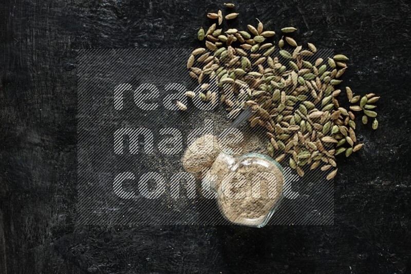 A flipped glass spice jar and a metal spoon full of cardamom powder and cardamom seeds spreaded on textured black flooring