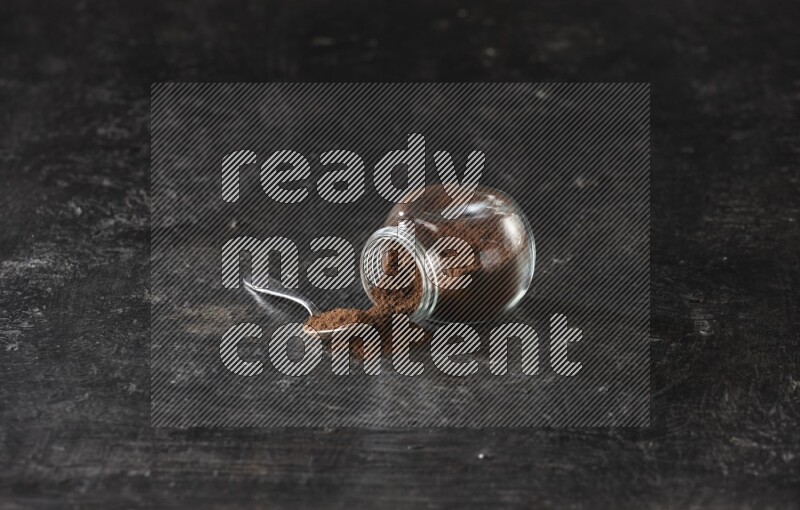 A flipped glass spice jar and a metal spoon full of cloves powder on textured black flooring