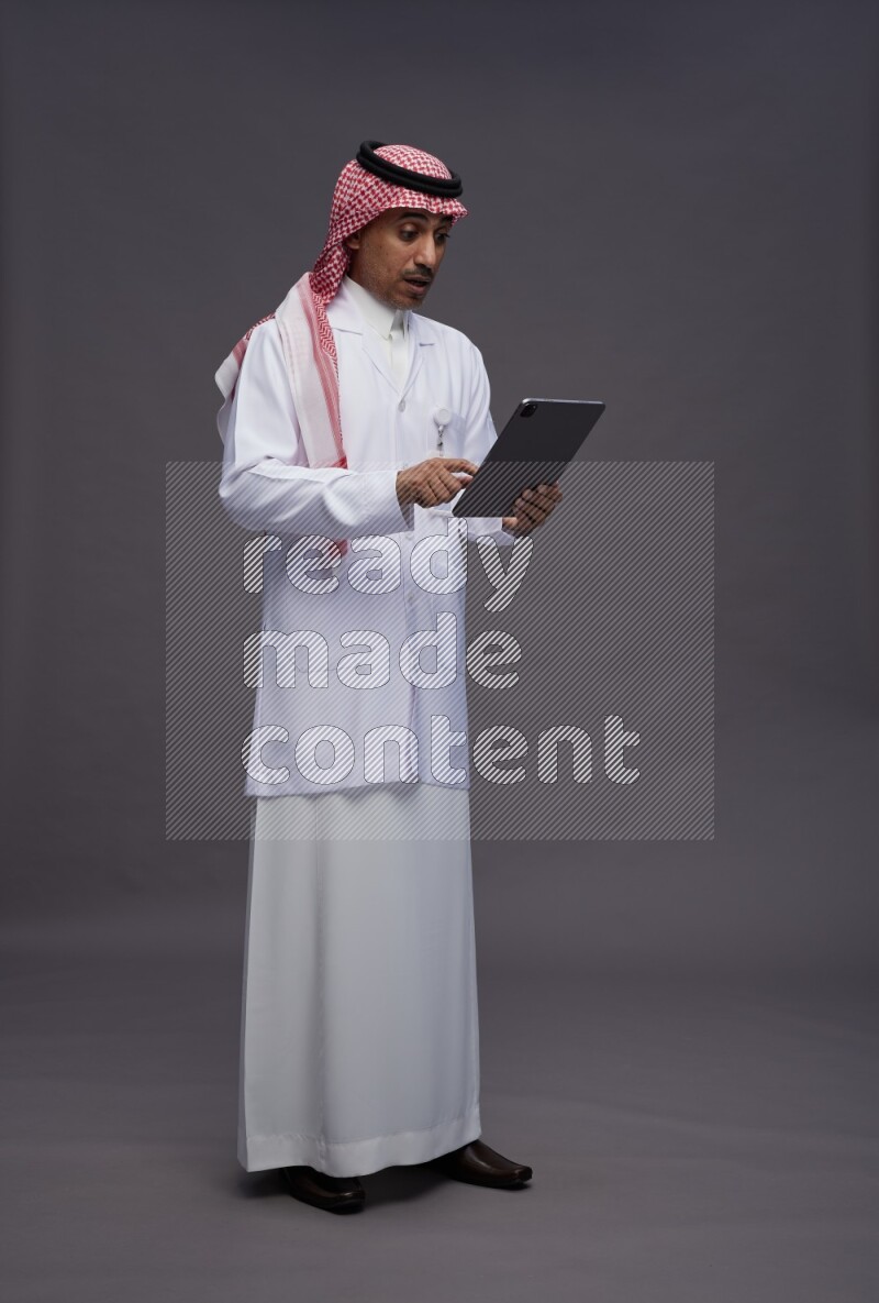 Saudi man wearing thob with lab coat and shomag with pocket employee badge standing working on tablet on gray background