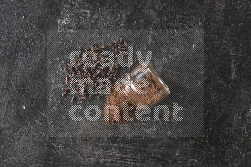 A flipped glass jar full of cloves powder with cloves spread on a textured black flooring