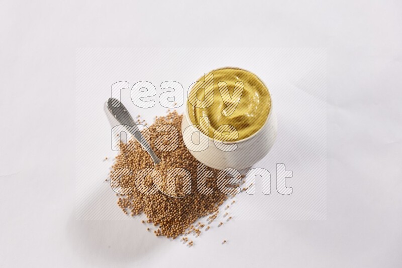 A beige pottery bowl full of mustard paste with mustard seeds underneath with a metal spoon on white flooring