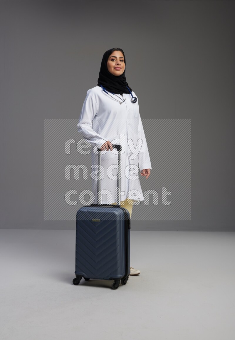 Saudi woman wearing lab coat with stethoscope standing holding Travel bag on Gray background