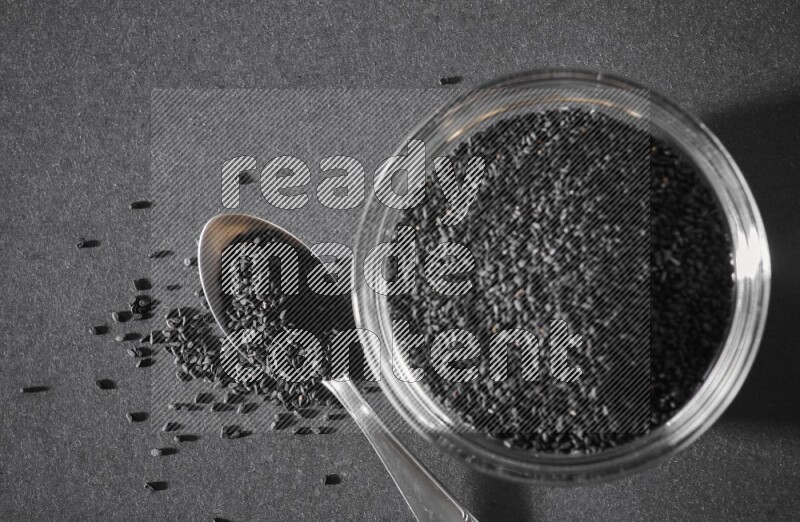 A glass bowl full of black seeds with a metal spoon full of the seeds on a black flooring