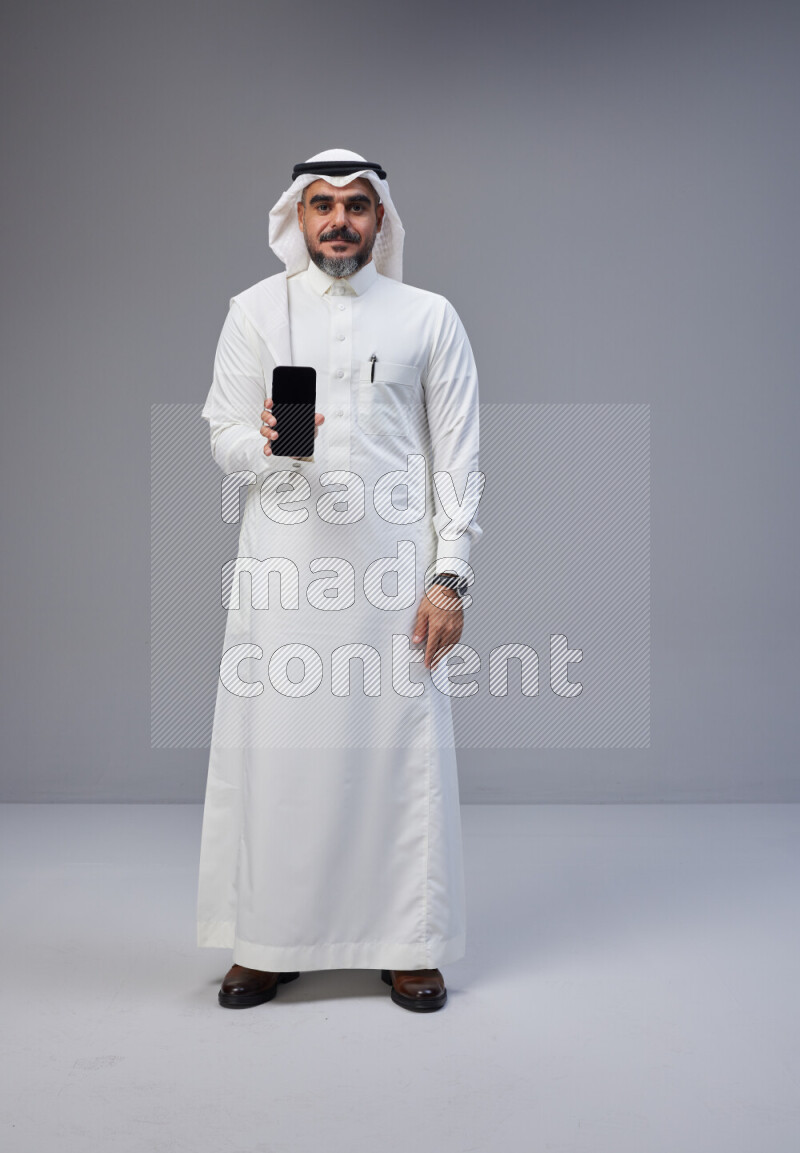 Saudi man Wearing Thob and white Shomag standing showing phone to camera on Gray background