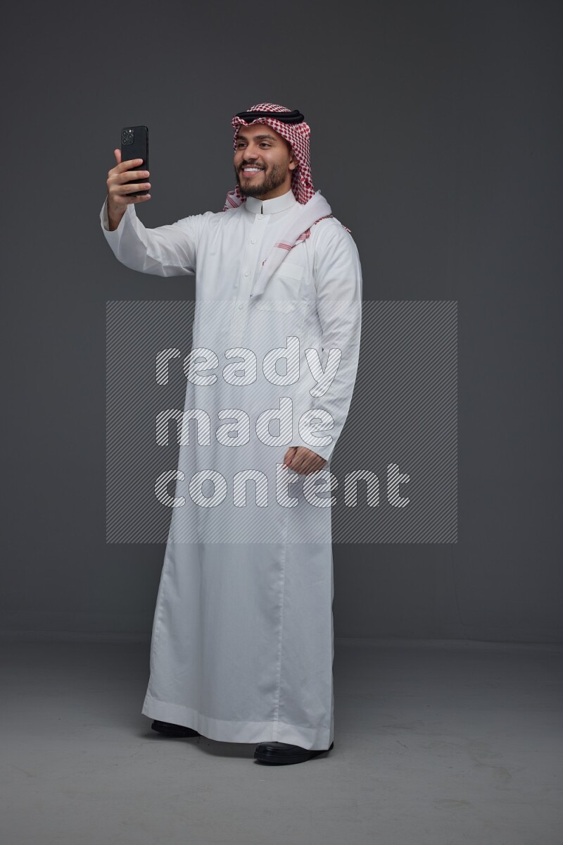 A Saudi man wearing Thobe and Shamgh taking selfie with his phone eye level on a gray background
