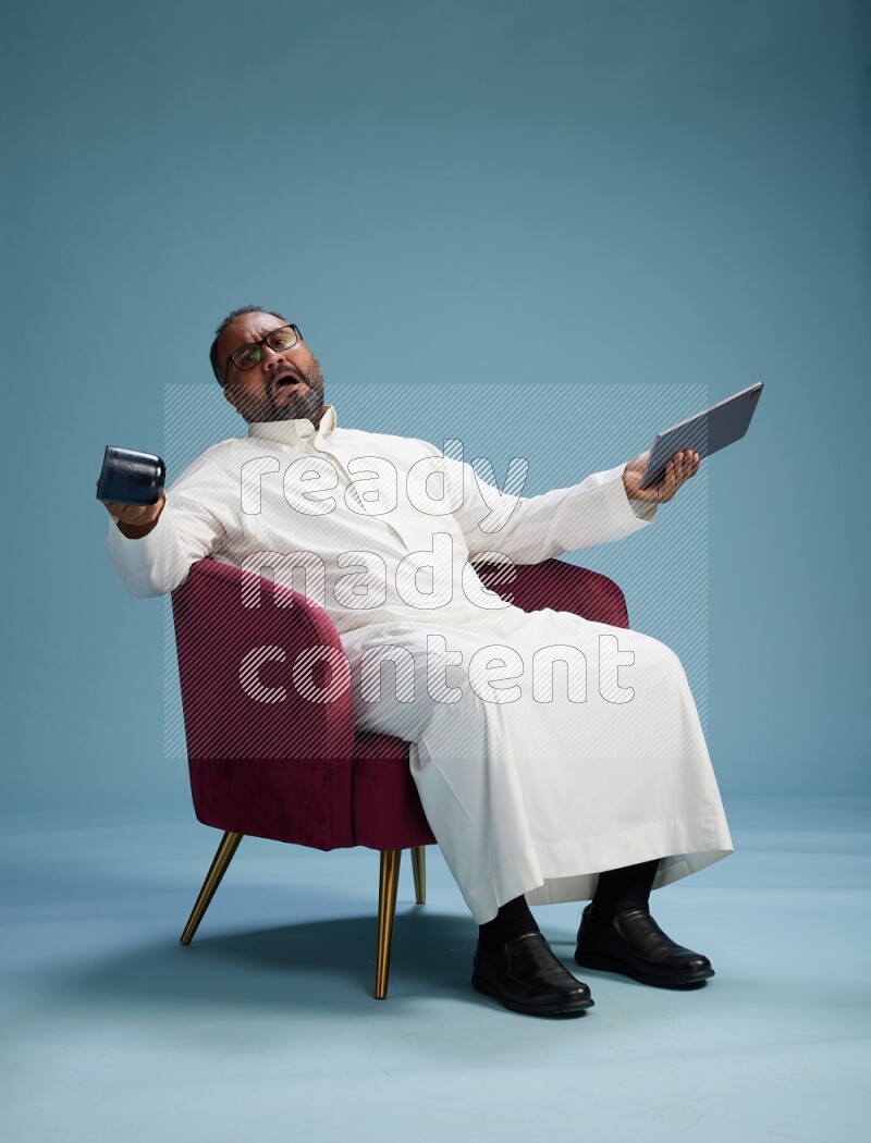 Saudi Man without shimag sitting on chair working on tablet on blue background