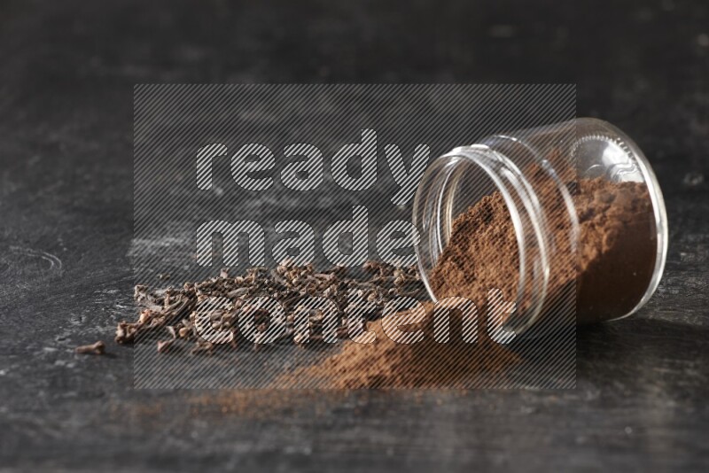 A flipped glass jar full of cloves powder with cloves spread on a textured black flooring