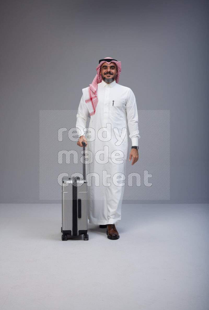 Saudi man wearing Thob and red Shomag standing holding Travel bag on Gray background