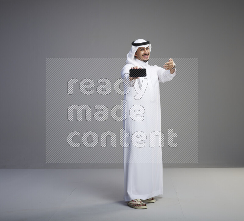 A Saudi man standing wearing thob and white shomag showing phone to camera on gray background