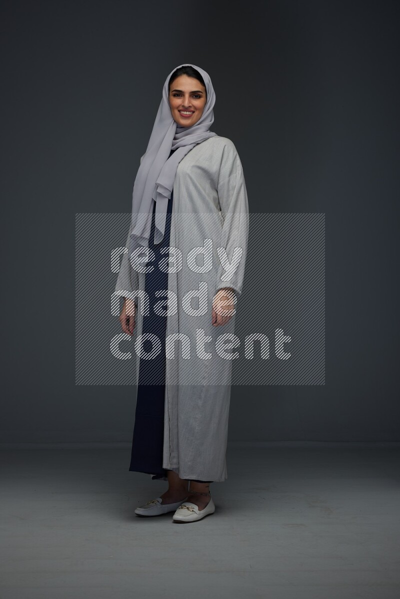 A Saudi woman wearing a light gray Abaya and head scarf standing and making multi poses eye level on a grey background