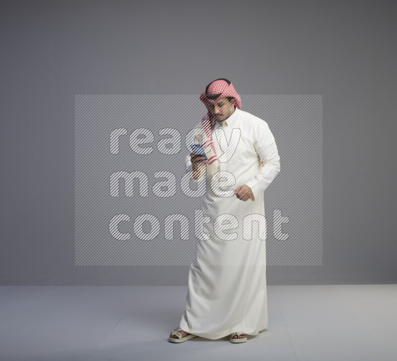 A  Saudi man standing wearing thob and red shomag texting on phone on gray background