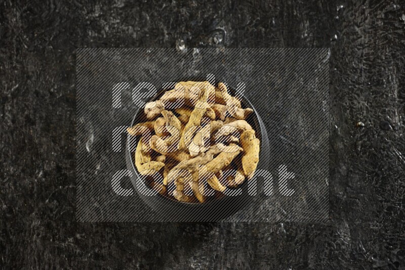 A black pottery bowl full of dried turmeric whole fingers on textured black flooring