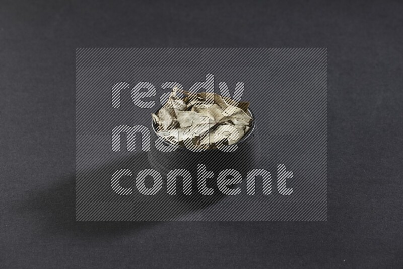 A black pottery bowl full of dried bay leaves on black flooring