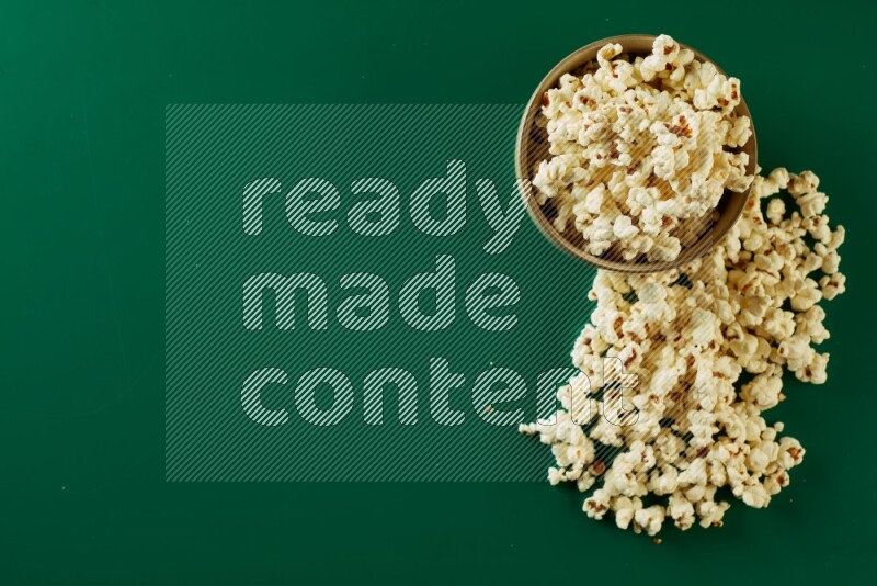 An off white ceramic bowl full of popcorn with popcorn beside it on a green background in a top view shot