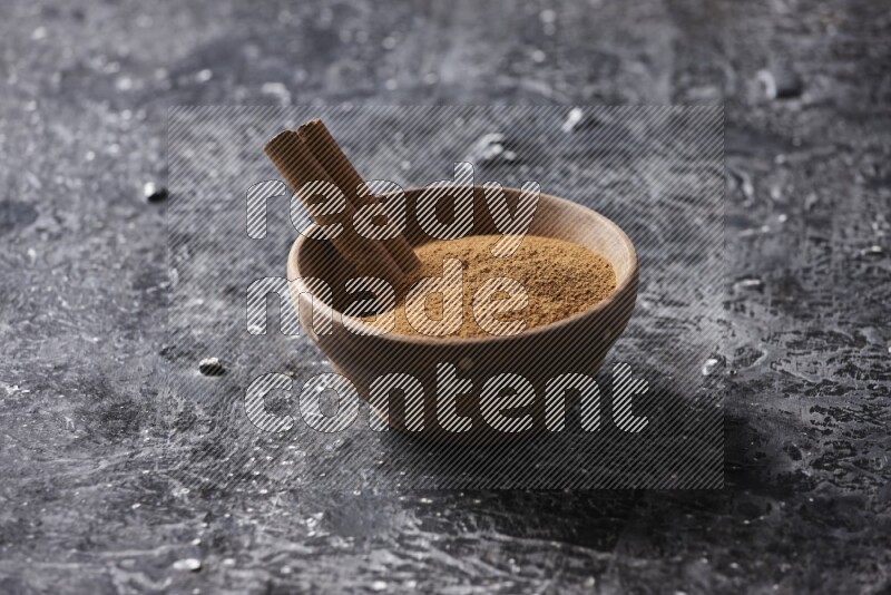 Wooden bowl full of cinnamon powder and a cinnamon stick on a textured black background