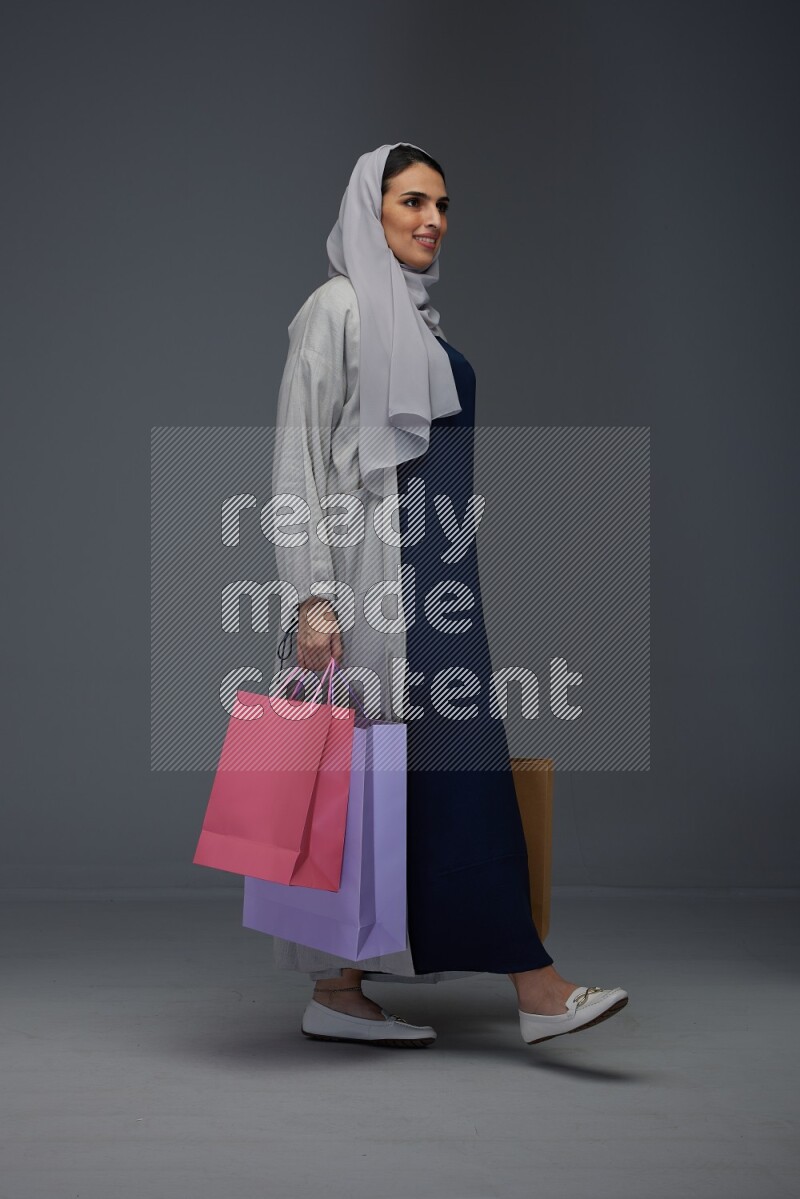 A Saudi woman wearing a light gray Abaya and head scarf standing and holding shopping bags on a grey background