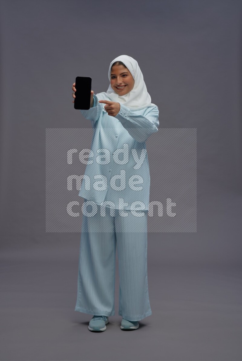 Saudi woman wearing hijab clothes standing showing phone to camera on gray background