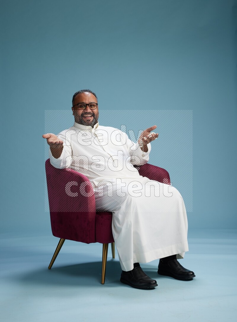 Saudi Man without shimag sitting on chair Interacting with the camera on blue background