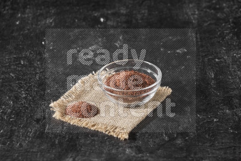 A glass bowl full of garden cress seeds with bunch of the seeds on burlap fabric on a textured black flooring
