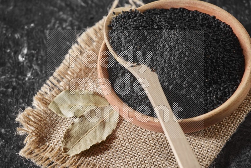 A wooden bowl full of black seeds with wooden spoon full of the seeds on it on a burlap fabric on a textured black flooring