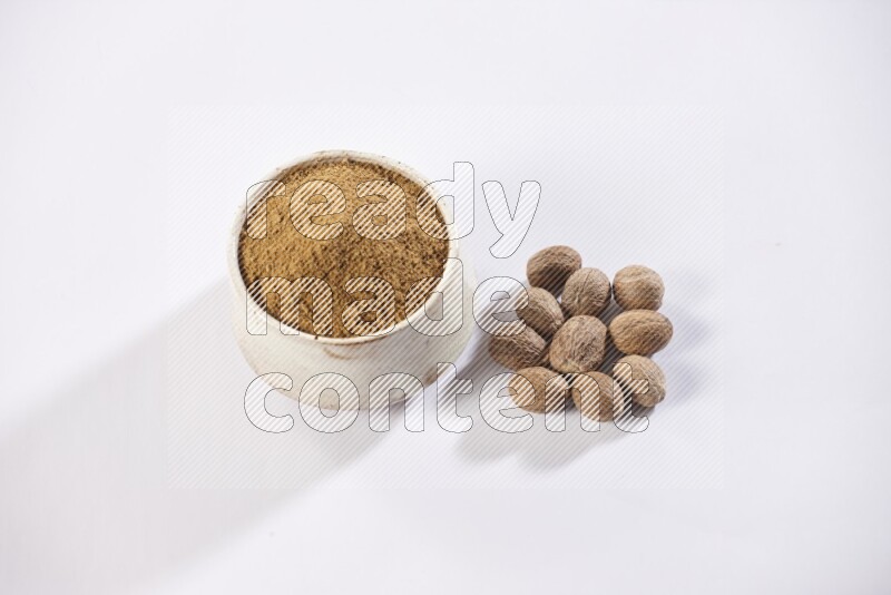 A beige pottery bowl full of nutmeg powder with whole seeds beside it on a white flooring