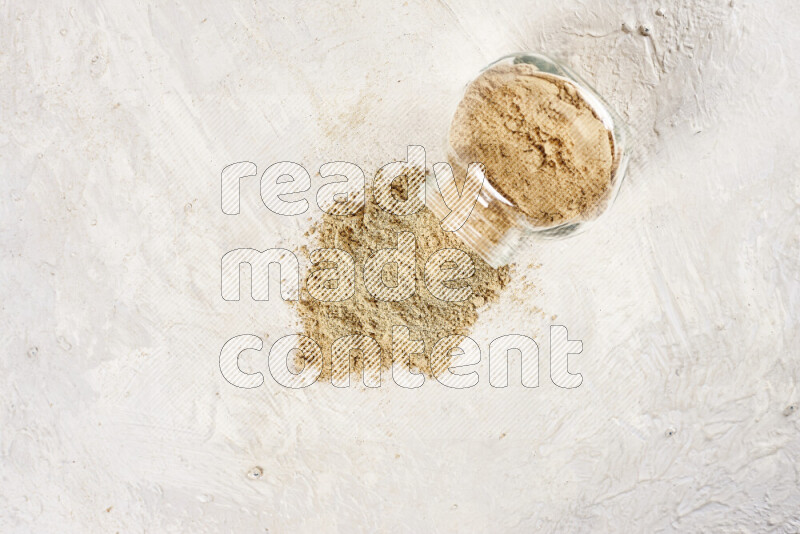 A glass jar full of ground ginger powder flipped with some spilling powder on white background